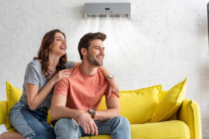 Cheerful couple enjoying comfort on a yellow sofa under an air conditioning unit, emphasizing the importance of HVAC services for home comfort in humid summer conditions.